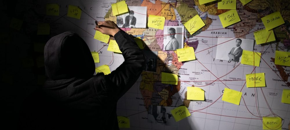 Back view of man planning hacker attack in dark room. Man writing on wall with stickers, photographs and red threads. Planning, conspiracy, hacking concept