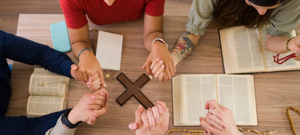 High angle of catholic young men and women holding each other hands while praying together around a table with a christian cross