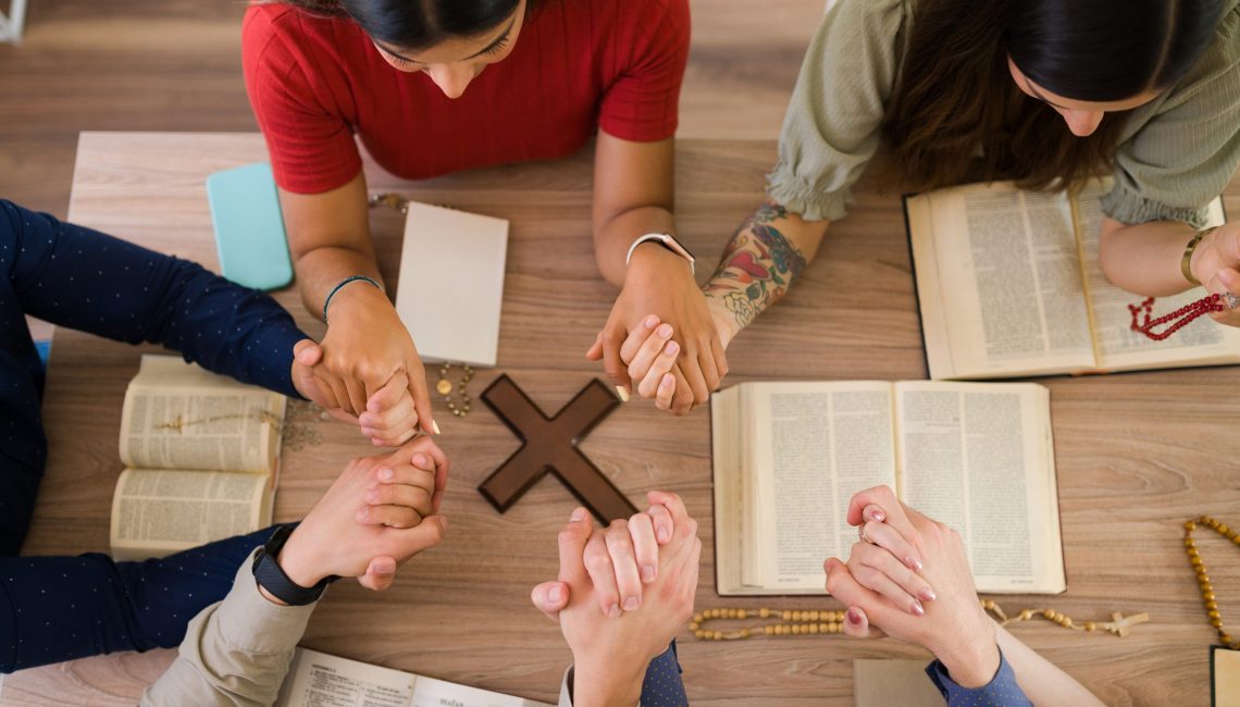 High angle of catholic young men and women holding each other hands while praying together around a table with a christian cross